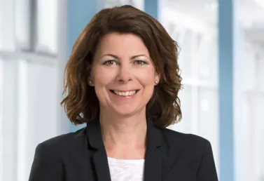 Frau mit lockigem, braunem Haar, lächelnd, trägt einen schwarzen Blazer und ein weißes Oberteil, vor einem hellen, modernen Hintergrund.