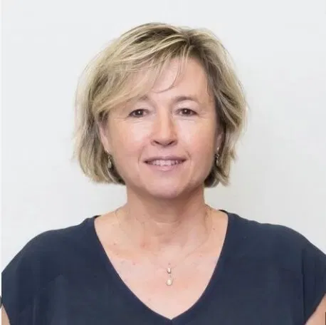 Porträt einer lächelnden Frau mit kurzem blondem Haar, dunklem Oberteil und zarter Halskette vor hellem Hintergrund.