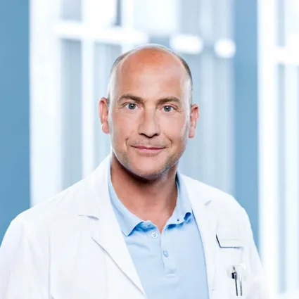 Mann mit Glatze und freundlichem Gesichtsausdruck, trägt einen weißen Laborkittel über einem hellblauen Poloshirt. Im Hintergrund sind unscharfe Fenster und helle Wände zu sehen, die eine klinische Umgebung andeuten.