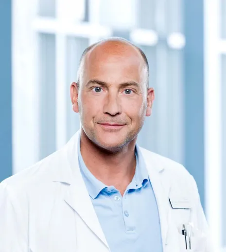 Mann mit Glatze und freundlichem Gesichtsausdruck, trägt einen weißen Laborkittel über einem hellblauen Poloshirt. Im Hintergrund sind unscharfe Fenster und helle Wände zu sehen, die eine klinische Umgebung andeuten.