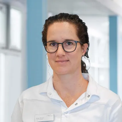 Frau mit lockigem, braunem Haar und Brille, trägt ein weißes Oberteil. Im Hintergrund sind helle Wände und Fenster zu sehen.