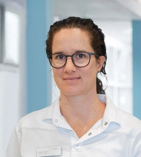 Frau mit lockigem, braunem Haar und Brille, trägt ein weißes Oberteil. Im Hintergrund sind helle Wände und Fenster zu sehen.