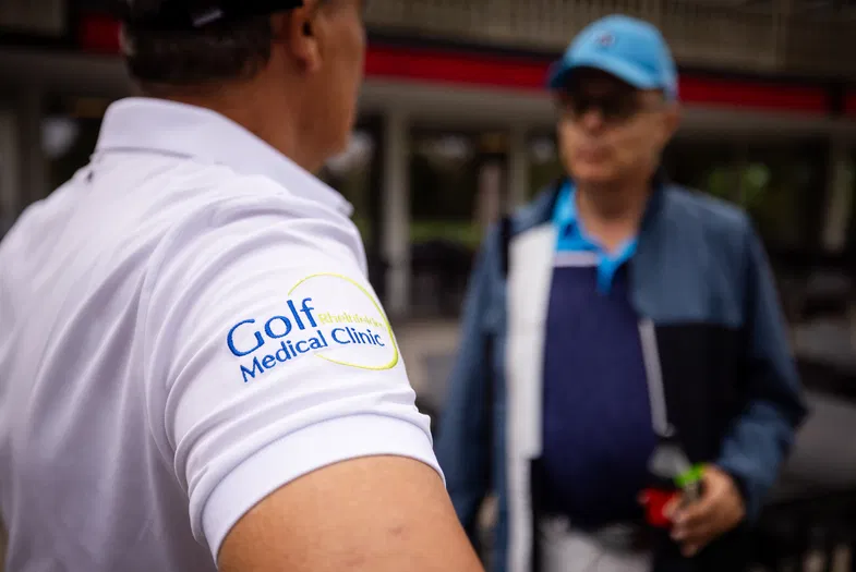 Nahaufnahme eines Golfspielers mit Logo „Golf Medical Clinic“ auf dem weißen Poloshirt; im Hintergrund eine unscharfe Person in Golfkleidung.