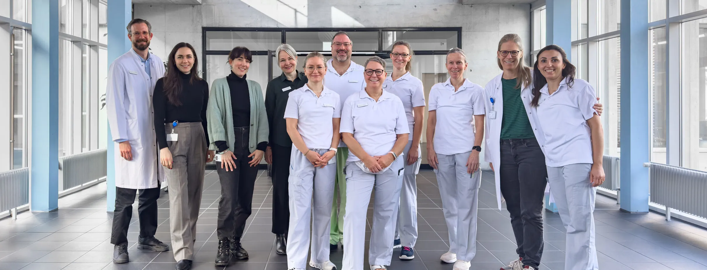 Gruppe von zwölf Personen in einem modernen Innenraum mit großen Fenstern, darunter medizinisches Personal in weißen Uniformen und einige in zivil. Sie stehen lächelnd zusammen, während ein Arzt in einem weißen Kittel links steht. Der Boden ist geflies...