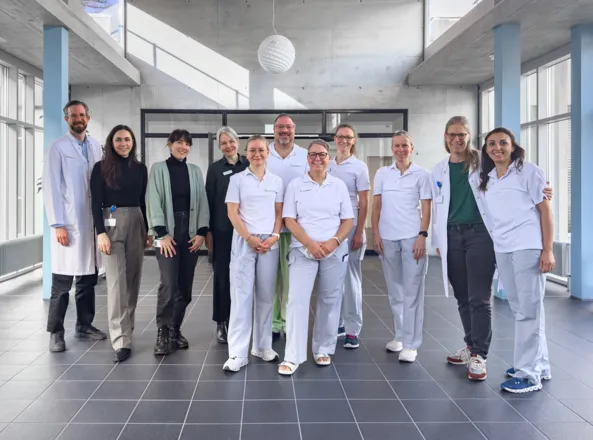 Gruppe von zwölf Personen in einem modernen Innenraum mit großen Fenstern, darunter medizinisches Personal in weißen Uniformen und einige in zivil. Sie stehen lächelnd zusammen, während ein Arzt in einem weißen Kittel links steht. Der Boden ist geflies...