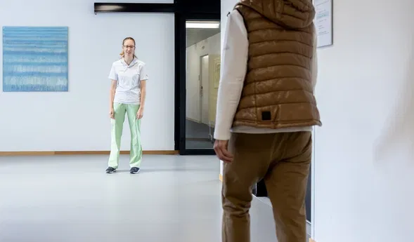 Krankenhausflur mit der Zimmernummer 2; eine Pflegekraft steht einer Patientin in brauner Weste gegenüber.