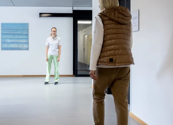 Krankenhausflur mit der Zimmernummer 2; eine Pflegekraft steht einer Patientin in brauner Weste gegenüber.
