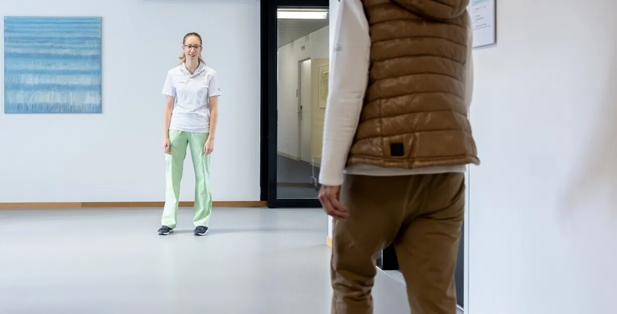 Krankenhausflur mit der Zimmernummer 2; eine Pflegekraft steht einer Patientin in brauner Weste gegenüber.