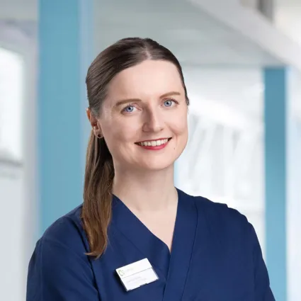 Frau mit langen, braunen Haaren und blauen Augen, trägt einen dunkelblauen Kittel und ein Namensschild. Heller, moderner Hintergrund mit blauen Akzenten.