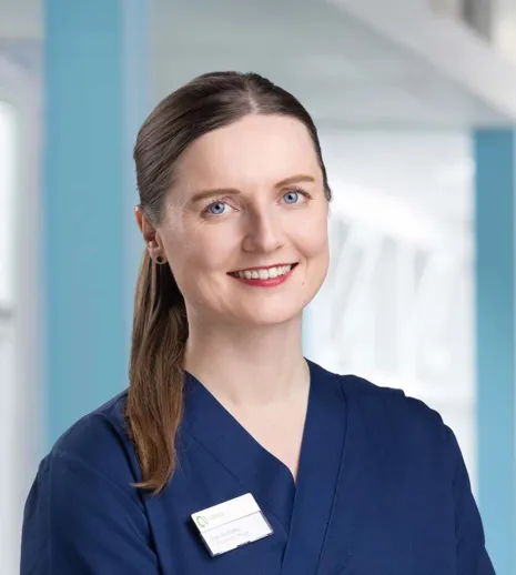 Frau mit langen, braunen Haaren und blauen Augen, trägt einen dunkelblauen Kittel und ein Namensschild. Heller, moderner Hintergrund mit blauen Akzenten.