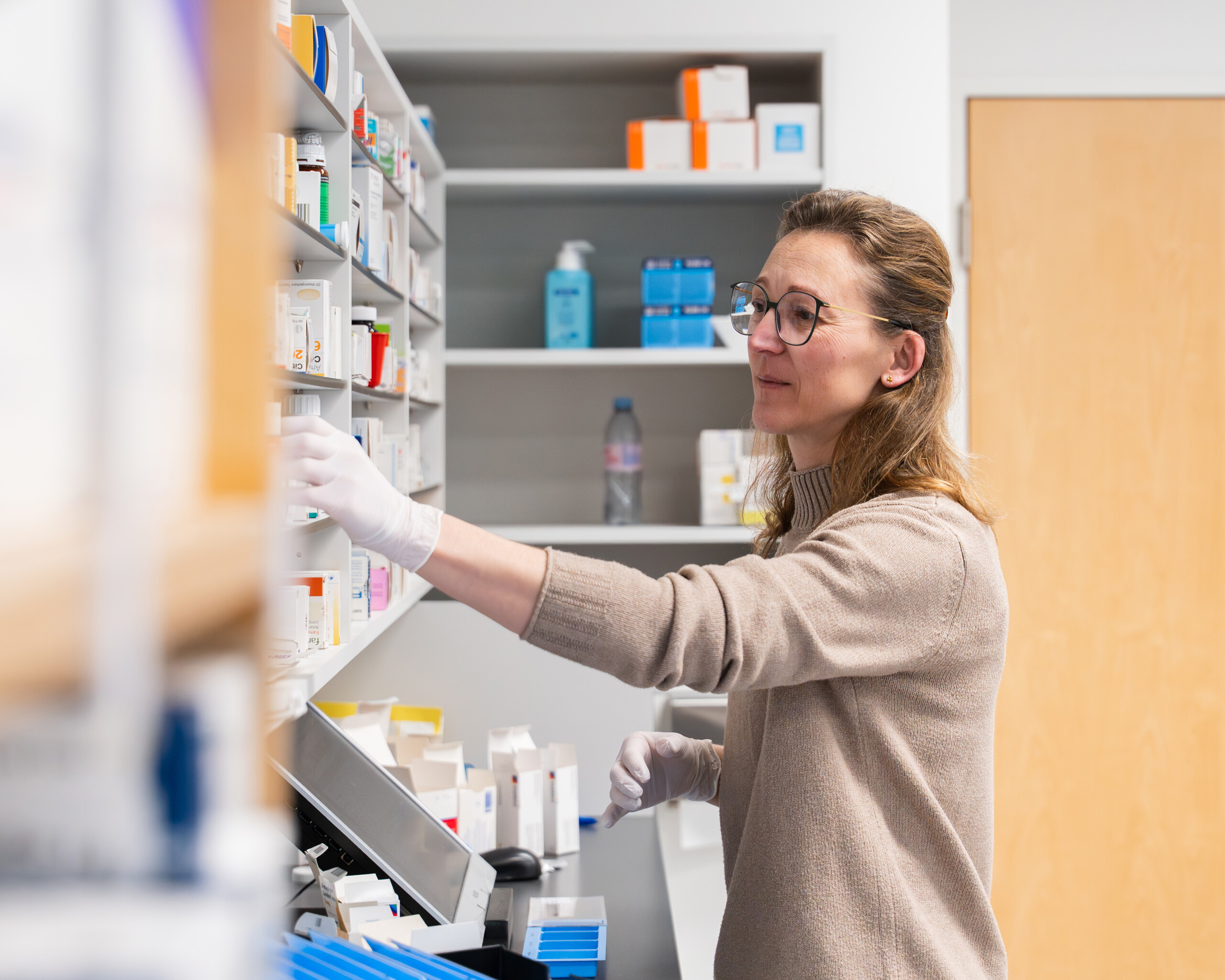 Apothekerin mit Brille und Handschuhen entnimmt Medikamente aus einem Regal in einer Apotheke.
