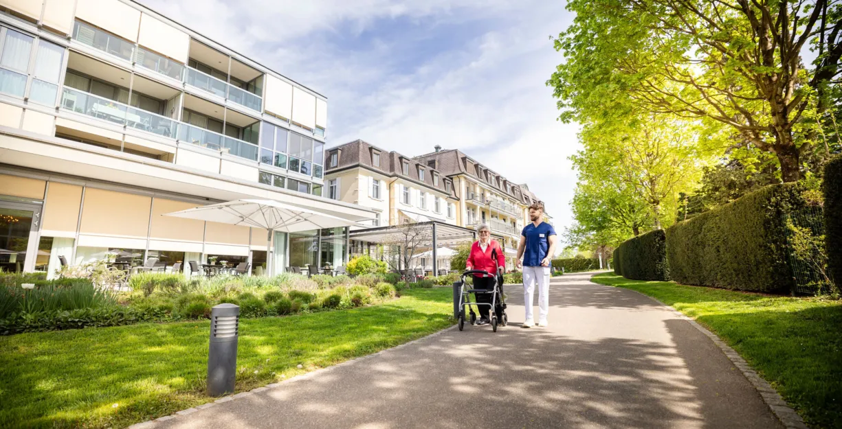Fußweg zwischen gepflegtem Grün und Hecken entlang eines modernen Wohn- und Bürogebäudes; zwei Personen gehen mit Fahrrad, sonniger Tag, Bäume und Schatten im Vordergrund.