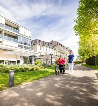 Fußweg zwischen gepflegtem Grün und Hecken entlang eines modernen Wohn- und Bürogebäudes; zwei Personen gehen mit Fahrrad, sonniger Tag, Bäume und Schatten im Vordergrund.