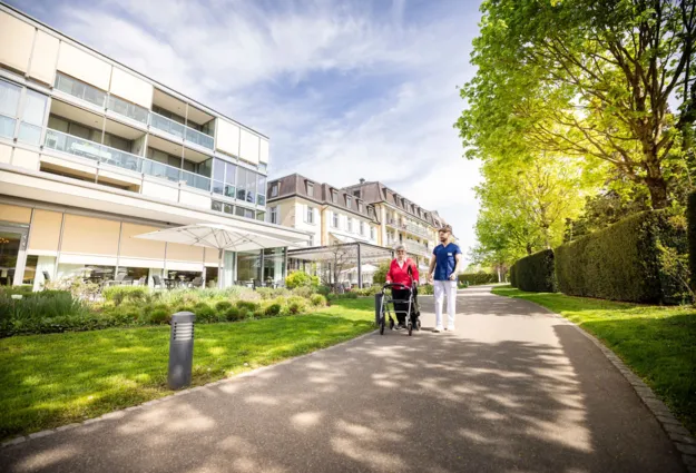 Fußweg zwischen gepflegtem Grün und Hecken entlang eines modernen Wohn- und Bürogebäudes; zwei Personen gehen mit Fahrrad, sonniger Tag, Bäume und Schatten im Vordergrund.