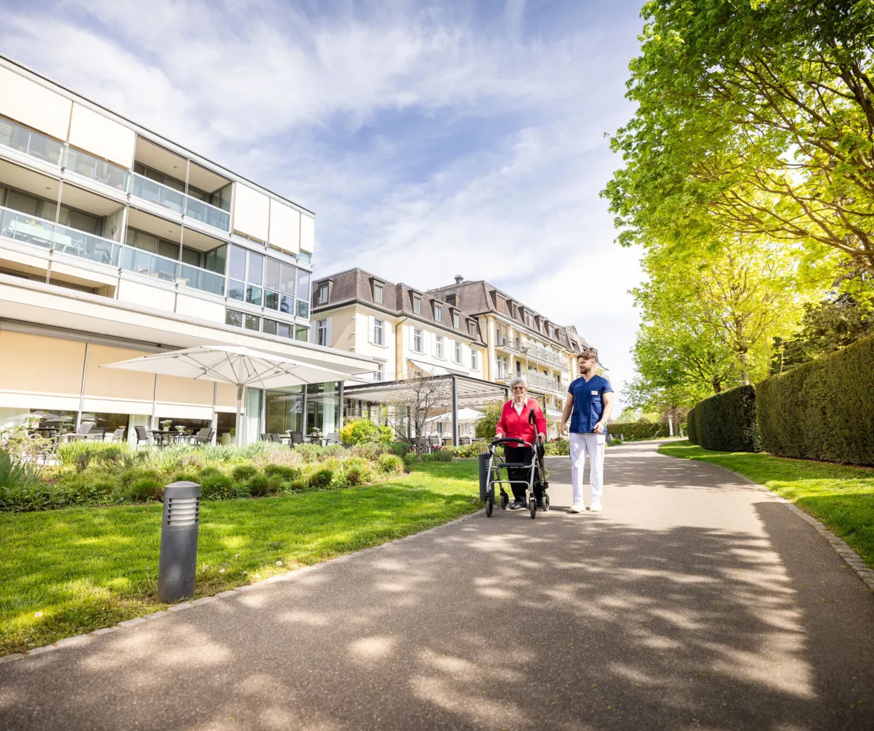 Fußweg zwischen gepflegtem Grün und Hecken entlang eines modernen Wohn- und Bürogebäudes; zwei Personen gehen mit Fahrrad, sonniger Tag, Bäume und Schatten im Vordergrund.