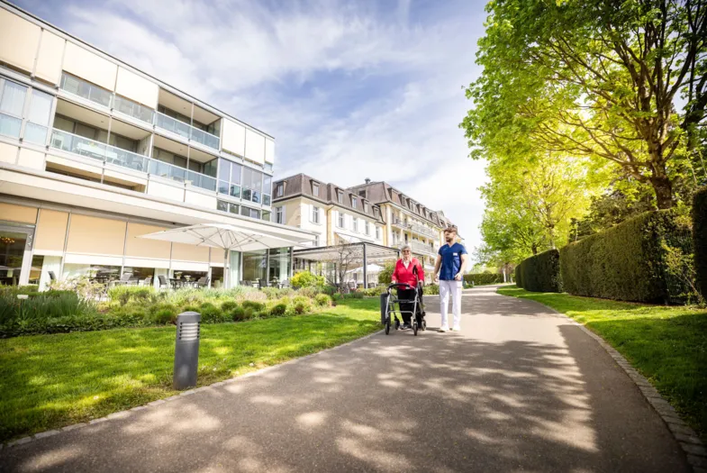 Fußweg zwischen gepflegtem Grün und Hecken entlang eines modernen Wohn- und Bürogebäudes; zwei Personen gehen mit Fahrrad, sonniger Tag, Bäume und Schatten im Vordergrund.
