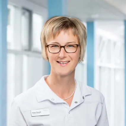 Frau mit blonden, kurzen Haaren und Brille, trägt ein weißes Oberteil mit Namensschild, lächelt vor einem hellen, modernen Hintergrund mit Fenstern.