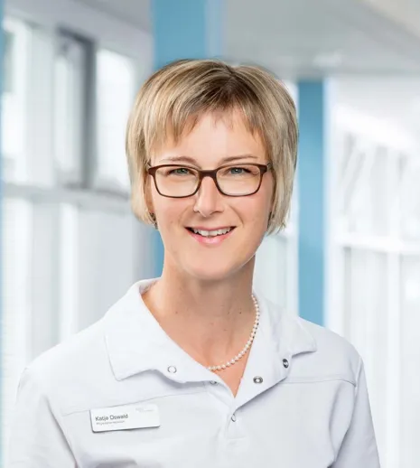 Frau mit blonden, kurzen Haaren und Brille, trägt ein weißes Oberteil mit Namensschild, lächelt vor einem hellen, modernen Hintergrund mit Fenstern.