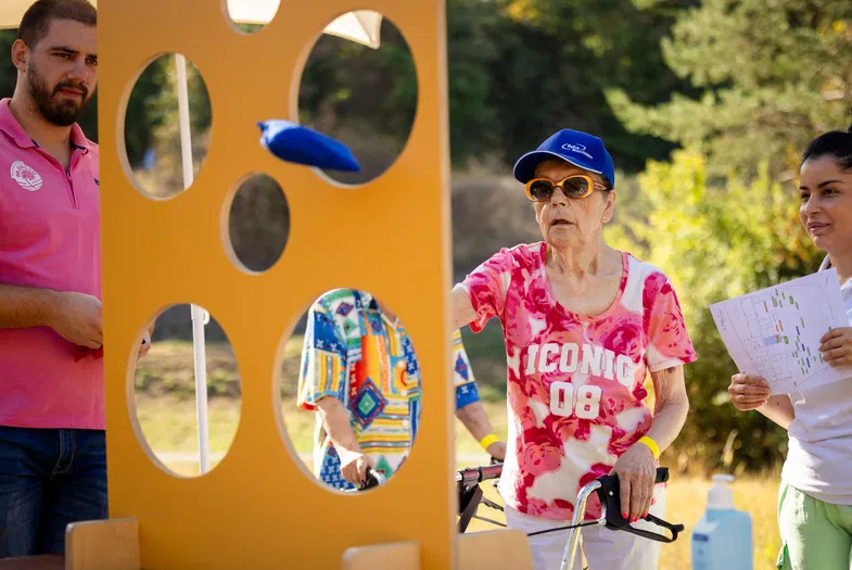Ältere Frau mit Rollator, pinkem Shirt und Sonnenbrille neben orangefarbener Spielstation; weitere Personen und eine Frau mit Plan im Freien.