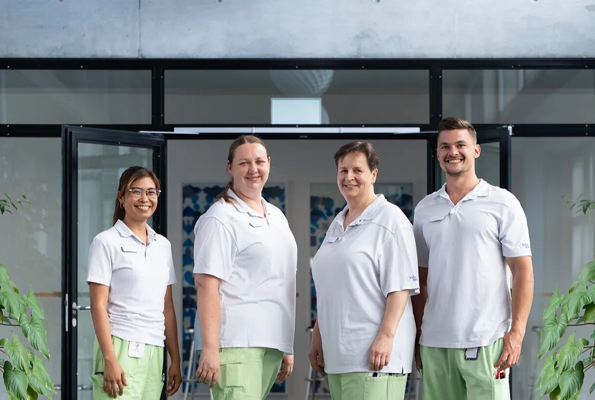 Vier Personen in weißen T-Shirts und grünen Hosen stehen vor einem Gebäude mit großen Glasfronten; links und rechts flankieren Pflanzen das Eingangsportal.