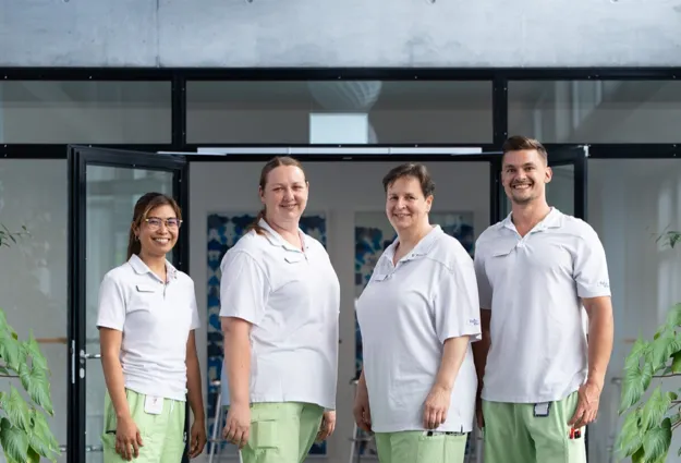 Vier Personen in weißen T-Shirts und grünen Hosen stehen vor einem Gebäude mit großen Glasfronten; links und rechts flankieren Pflanzen das Eingangsportal.