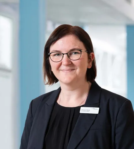 Porträt einer Frau mit dunkelbraunem kurzen Haar und Brille im Business-Look, schwarzer Blazer mit Namensschild, vor blau-weiß unscharfem Bürohintergrund.