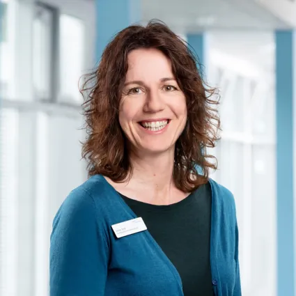 Lächelnde Frau mit lockigem, braunem Haar, trägt einen blauen Pullover über einem schwarzen Oberteil. Sie steht vor einem hellen, modernen Hintergrund mit großen Fenstern und trägt ein Namensschild.