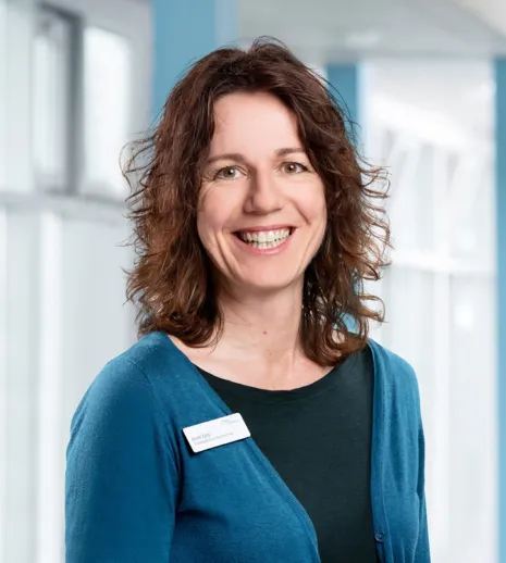 Lächelnde Frau mit lockigem, braunem Haar, trägt einen blauen Pullover über einem schwarzen Oberteil. Sie steht vor einem hellen, modernen Hintergrund mit großen Fenstern und trägt ein Namensschild.