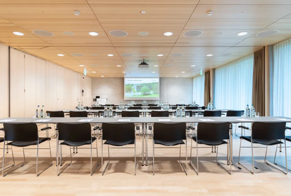 Konferenzraum mit mehreren Tischen und Stühlen, ausgestattet mit Wasserflaschen und Notizblättern. An der Wand ein Projektor mit einer Präsentation, große Fenster mit Vorhängen und Holzdecke.