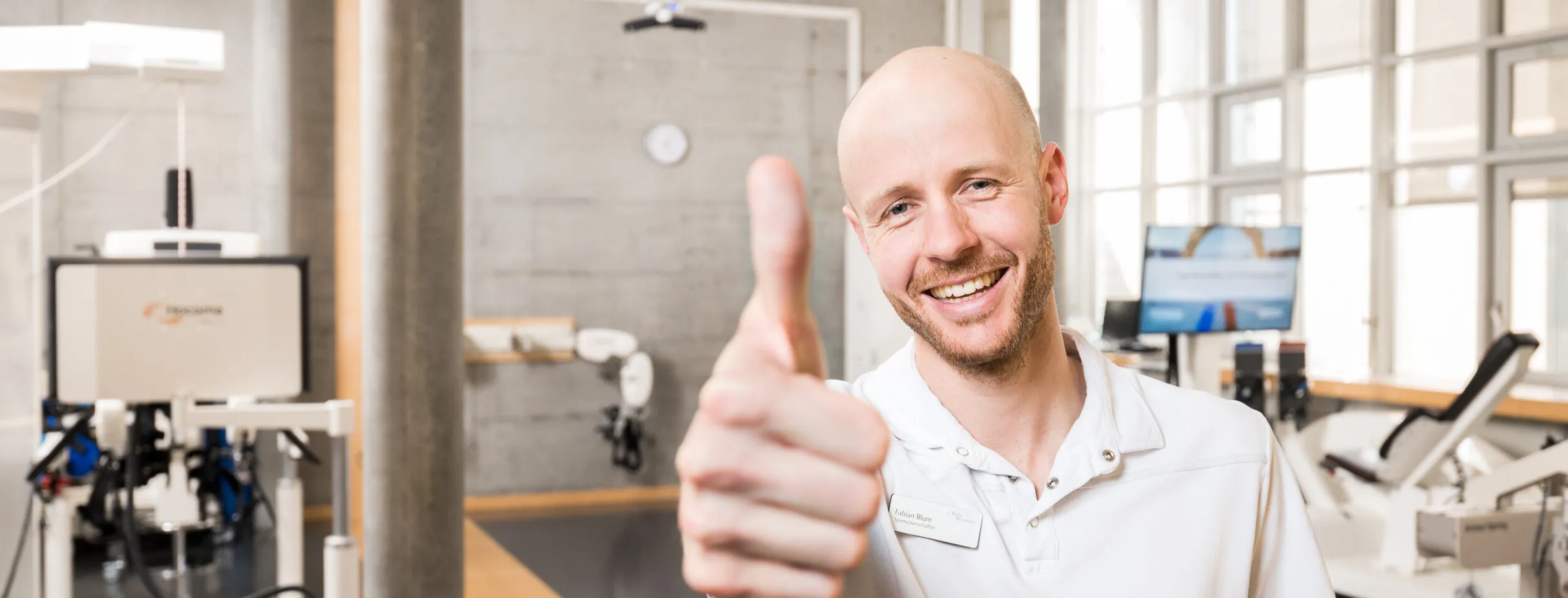 Lächelnder Mann mit Glatze und Bart zeigt Daumen hoch, trägt ein weißes Polohemd. Im Hintergrund sind moderne Trainingsgeräte und große Fenster sichtbar.