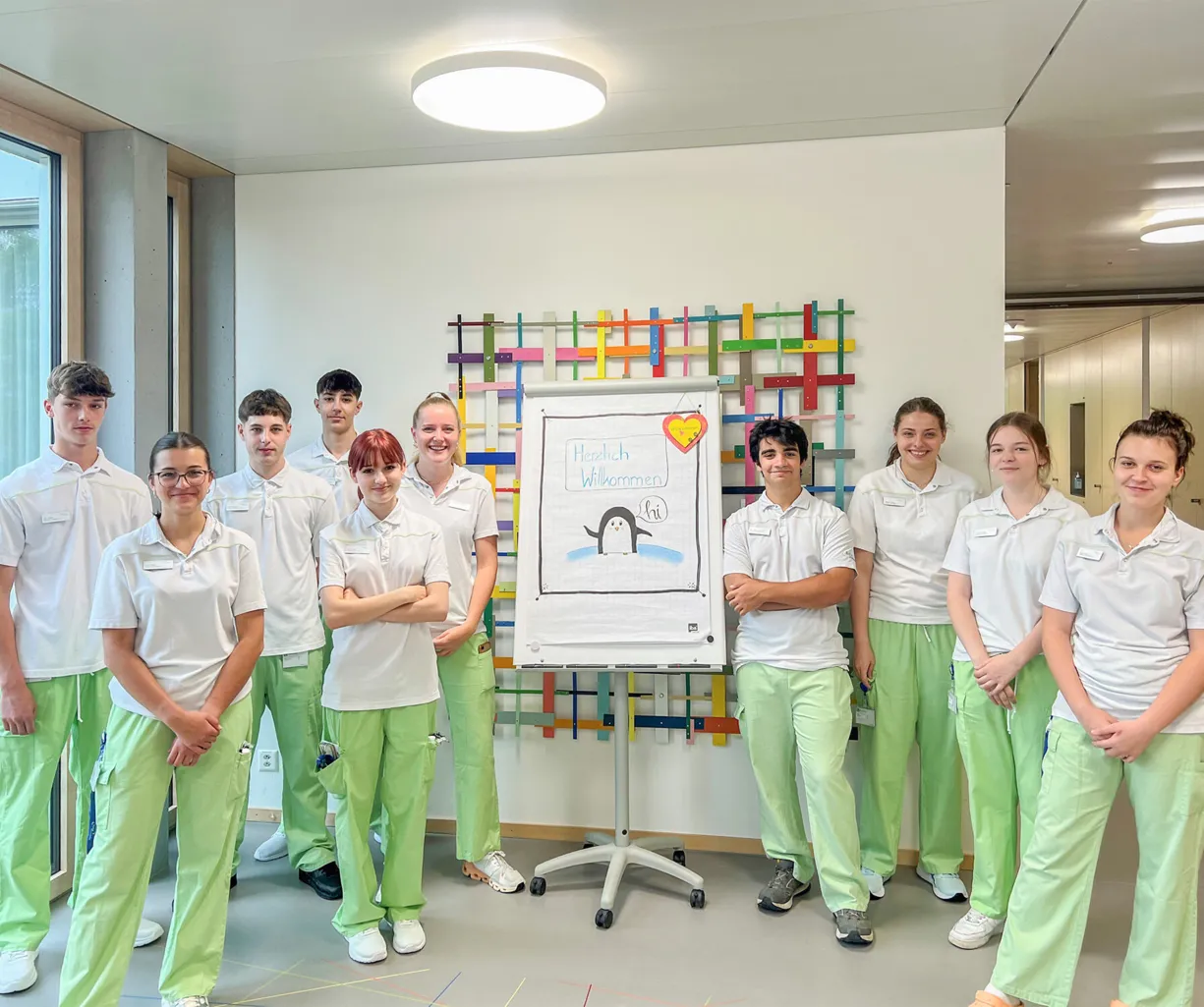 Gruppe von zehn Personen in einheitlicher Kleidung, bestehend aus weißen Oberteilen und hellgrünen Hosen, stehen vor einem Flipchart mit der Aufschrift „Herzlich Willkommen“ und einer Zeichnung eines Pinguins. Im Hintergrund sind große Fenster und eine...