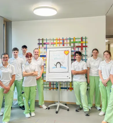 Gruppe von zehn Personen in einheitlicher Kleidung, bestehend aus weißen Oberteilen und hellgrünen Hosen, stehen vor einem Flipchart mit der Aufschrift „Herzlich Willkommen“ und einer Zeichnung eines Pinguins. Im Hintergrund sind große Fenster und eine...