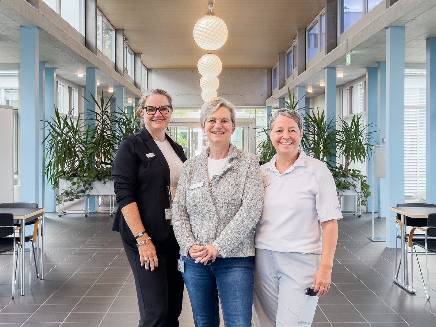 Drei Frauen stehen in einem modernen Innenraum mit großen Fenstern und Pflanzen. Die Frau links trägt einen schwarzen Blazer, die mittlere eine graue Strickjacke, und die Frau rechts ein weißes Polo-Shirt. Im Hintergrund sind Tische und Stühle sichtbar...