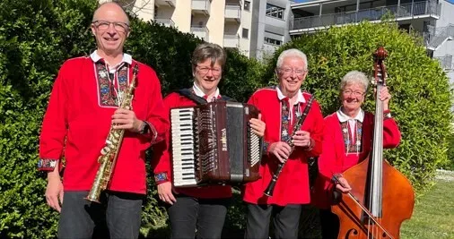 Vier Musiker in roten Jacken posieren im Freien mit Saxofon, Akkordeon, Klarinette und Kontrabass vor grünen Hecken vor einem Wohngebäude.