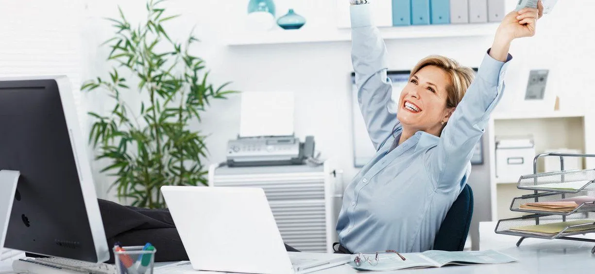 Frau mit erhobenen Händen sitzt entspannt am Schreibtisch, umgeben von Büroausstattung, einem Computer und Pflanzen. Sie lächelt und wirkt zufrieden.