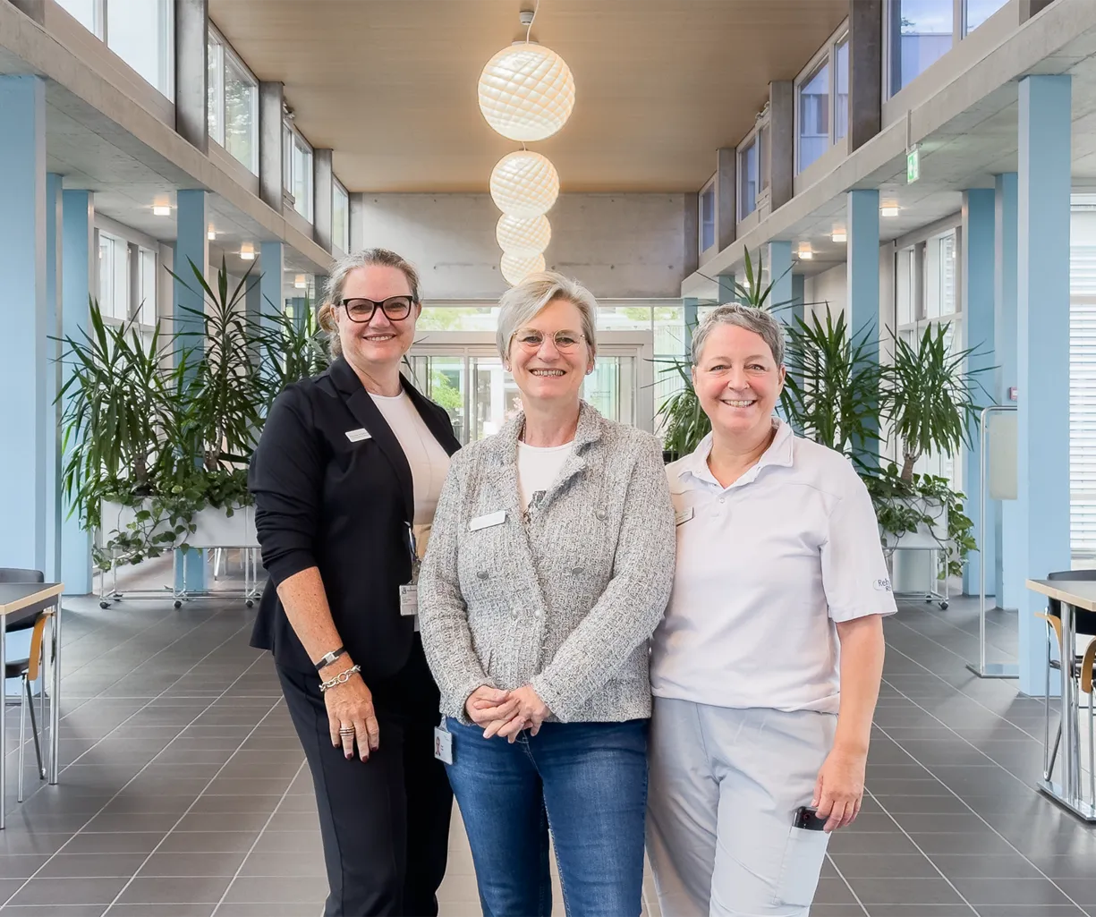 Drei Frauen stehen in einem modernen Innenraum mit großen Fenstern und Pflanzen. Die linke Frau trägt eine schwarze Blazerjacke, die mittlere eine graue Strickjacke und die rechte ein weißes Polo-Shirt. Im Hintergrund sind Tische und Stühle sichtbar.