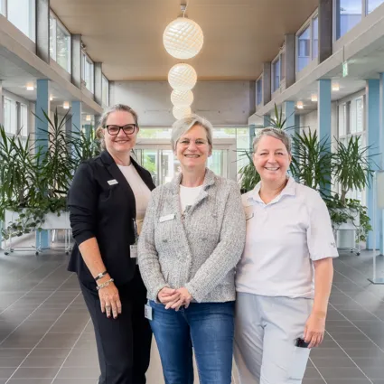 Drei Frauen stehen in einem modernen Innenraum mit großen Fenstern und Pflanzen. Die linke Frau trägt eine schwarze Blazerjacke, die mittlere eine graue Strickjacke und die rechte ein weißes Polo-Shirt. Im Hintergrund sind Tische und Stühle sichtbar.