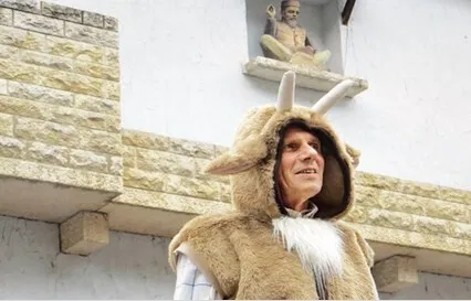 Mann in einem Fellkostüm mit Geweihkapuze und weißem Bart vor einer hellen Steinmauer; darüber sitzt eine kleine Heiligenfigur in einer Nische.