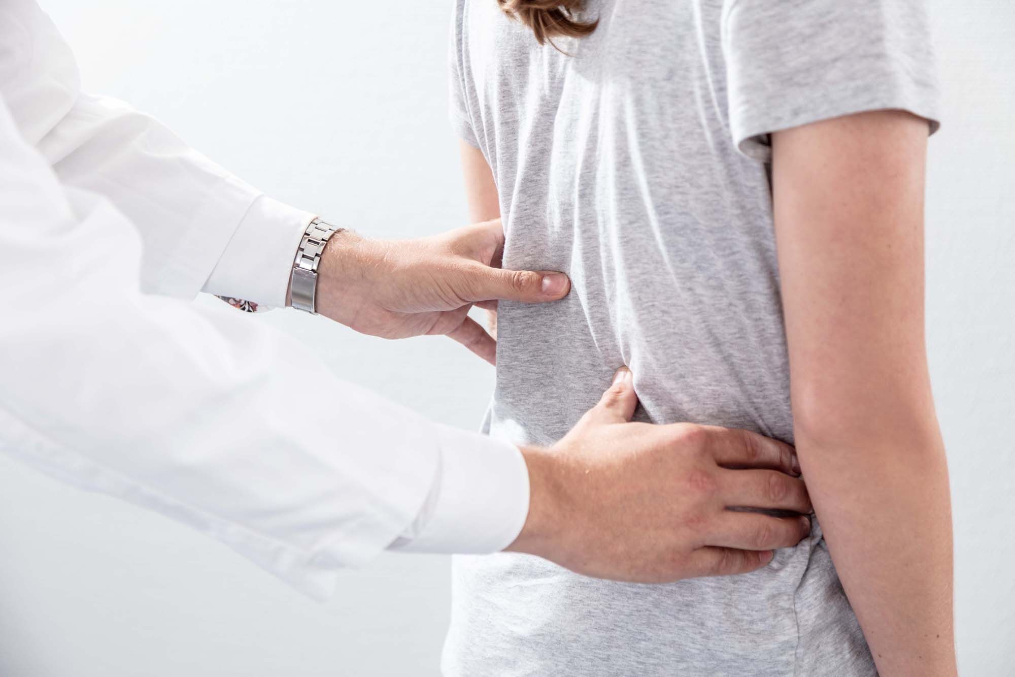 Hände eines Arztes, die den Rücken einer Person in einem grauen T-Shirt untersuchen, mit einem neutralen Hintergrund.