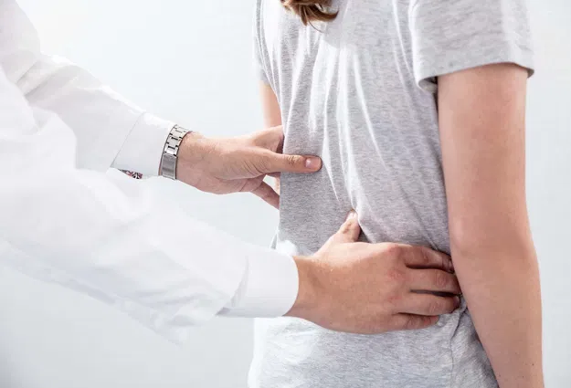 Hände eines Arztes, die den Rücken einer Person in einem grauen T-Shirt untersuchen, mit einem neutralen Hintergrund.