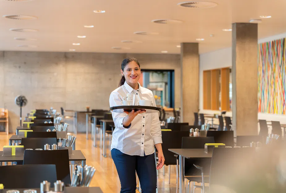 Lächelnde Kellnerin in weißem Hemd serviert ein Tablett in einem modernen, hellen Restaurant mit gedeckten Tischen und farbenfrohem Wandbild.