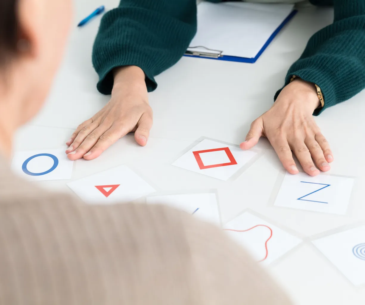 Zwei Personen an einem Tisch, eine Person von hinten sichtbar, die andere mit einer grünen Strickjacke. Auf dem Tisch liegen mehrere Karten mit geometrischen Formen und Buchstaben, darunter ein blauer Kreis, ein rotes Dreieck, ein rotes Quadrat und ein...