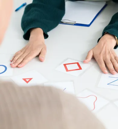 Zwei Personen an einem Tisch, eine Person von hinten sichtbar, die andere mit einer grünen Strickjacke. Auf dem Tisch liegen mehrere Karten mit geometrischen Formen und Buchstaben, darunter ein blauer Kreis, ein rotes Dreieck, ein rotes Quadrat und ein...