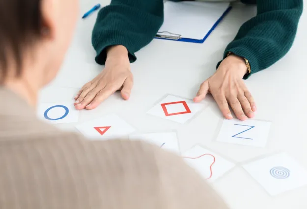 Zwei Personen an einem Tisch, eine Person von hinten sichtbar, die andere mit einer grünen Strickjacke. Auf dem Tisch liegen mehrere Karten mit geometrischen Formen und Buchstaben, darunter ein blauer Kreis, ein rotes Dreieck, ein rotes Quadrat und ein...