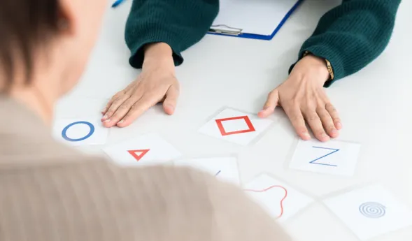 Zwei Personen an einem Tisch, eine Person von hinten sichtbar, die andere mit einer grünen Strickjacke. Auf dem Tisch liegen mehrere Karten mit geometrischen Formen und Buchstaben, darunter ein blauer Kreis, ein rotes Dreieck, ein rotes Quadrat und ein...