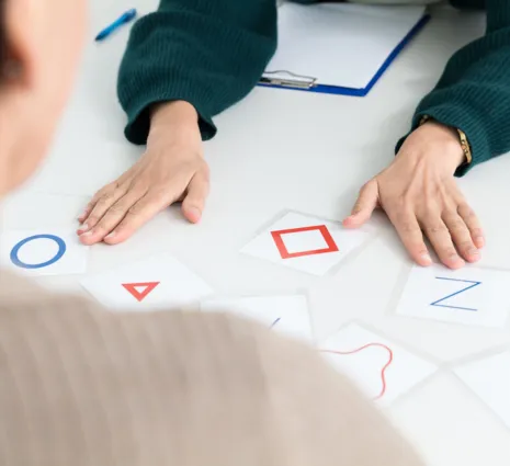 Zwei Personen an einem Tisch, eine Person von hinten sichtbar, die andere mit einer grünen Strickjacke. Auf dem Tisch liegen mehrere Karten mit geometrischen Formen und Buchstaben, darunter ein blauer Kreis, ein rotes Dreieck, ein rotes Quadrat und ein...