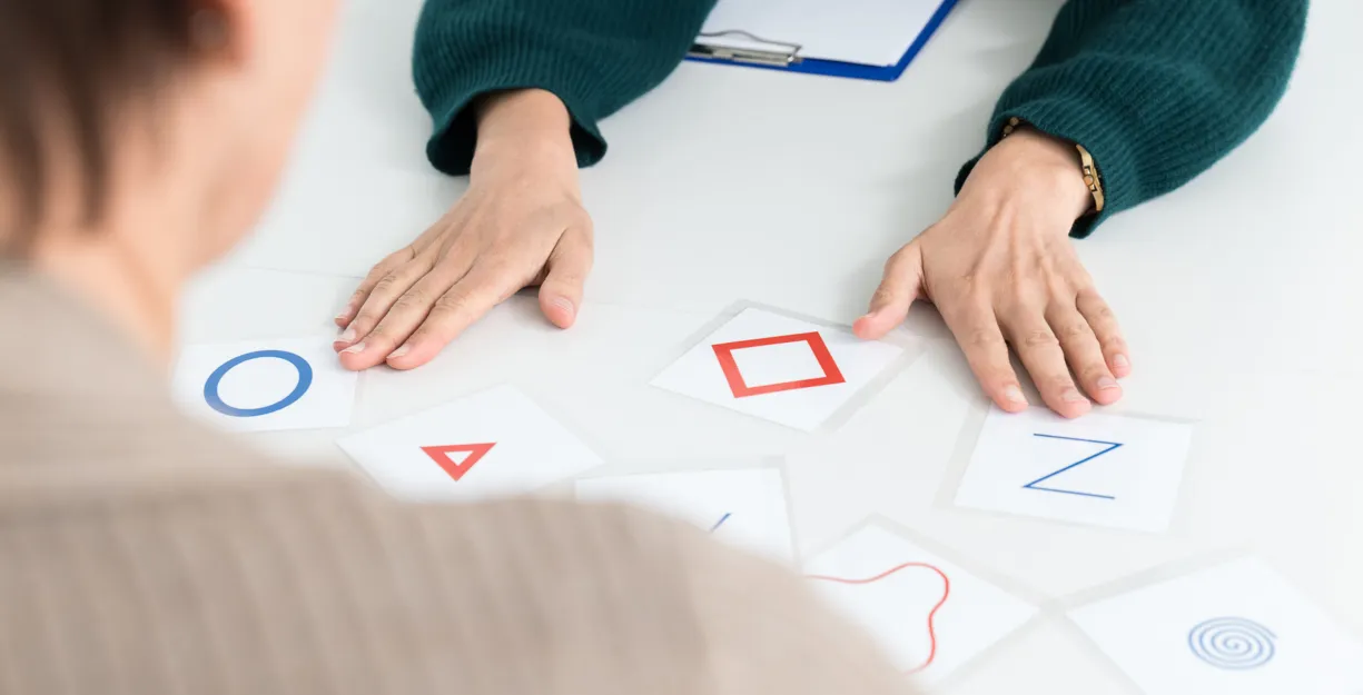 Zwei Personen an einem Tisch, eine Person von hinten sichtbar, die andere mit einer grünen Strickjacke. Auf dem Tisch liegen mehrere Karten mit geometrischen Formen und Buchstaben, darunter ein blauer Kreis, ein rotes Dreieck, ein rotes Quadrat und ein...