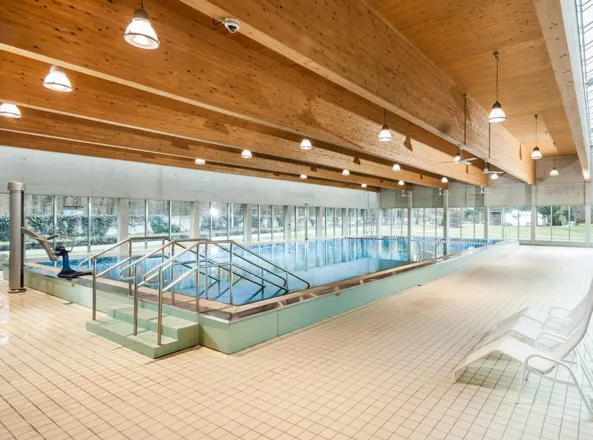 Innenbereich eines Schwimmbads mit einem großen, hellen Becken, umgeben von großen Fenstern. Holzdecke mit sichtbaren Balken, moderne Beleuchtung und Fliesenboden. An einer Seite eine Treppe und ein Handlauf, sowie einige Liegen in der Nähe des Beckens...