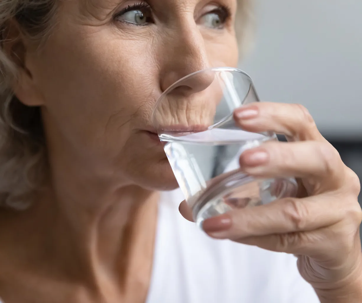 Ältere Frau trinkt aus einem Glas Wasser, Seitenansicht, Hände halten das transparente Trinkglas vor dem Mund, Fokus auf Gesicht und Hände.