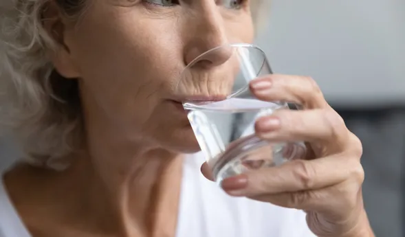 Ältere Frau trinkt aus einem Glas Wasser, Seitenansicht, Hände halten das transparente Trinkglas vor dem Mund, Fokus auf Gesicht und Hände.